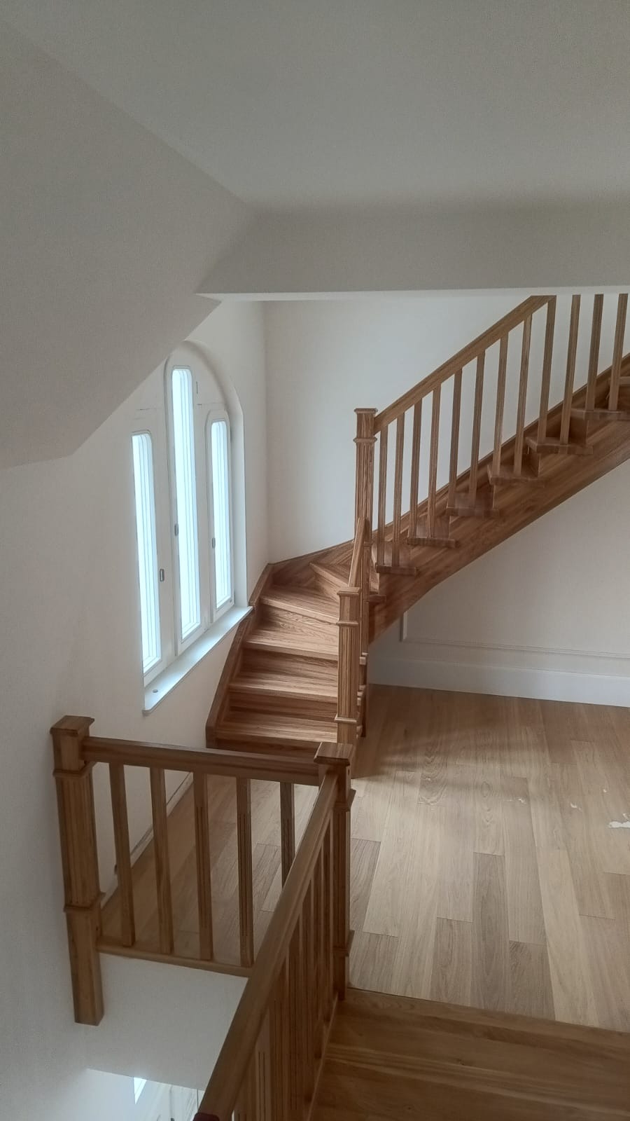 Jasne, drewniane schody z balustradą, zakręcające w lewo, widziane z góry. Obok okno z trzema pionowymi szybami. Podłoga z paneli w odcieniach beżu.