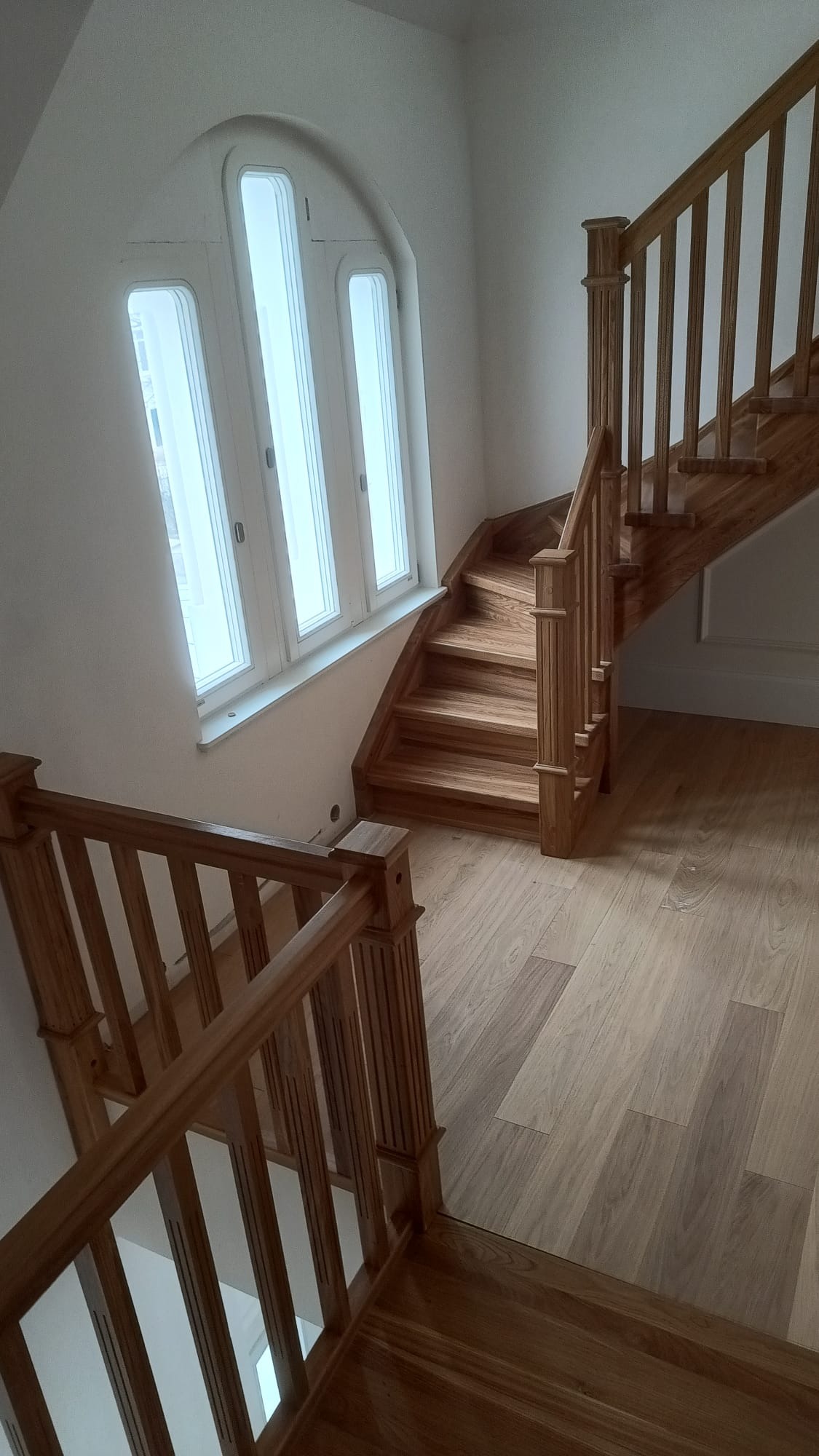 Jasne, drewniane schody z ozdobnymi balustradami, prowadzące w górę, obok okna łukowego. Parkiet na podłodze. Minimalistyczne wnętrze.