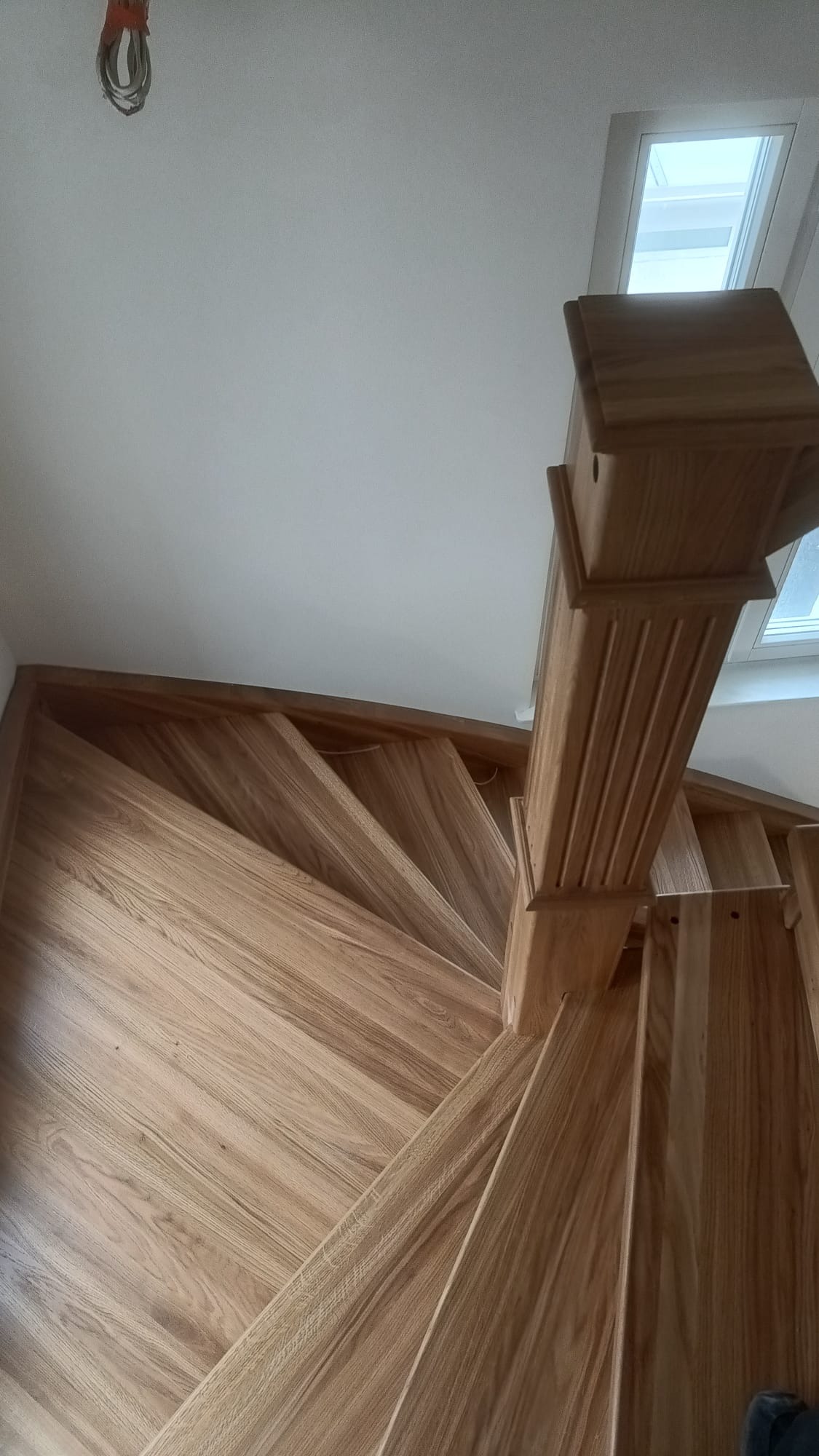 Drewniane schody kręcone z jasnego drewna, widok z dołu, z elementami dekoracyjnymi na słupku, oświetlone światłem z okna dachowego.