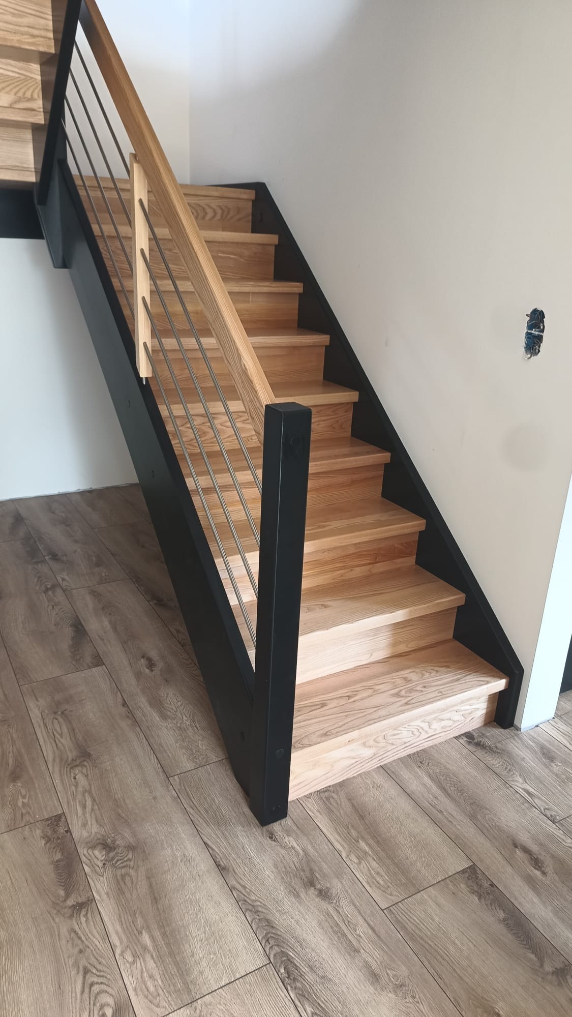 Drewniane schody z czarną metalową balustradą i drewnianą poręczą, widok z dołu, na podłodze panele imitujące drewno, gniazdko elektryczne na ścianie.