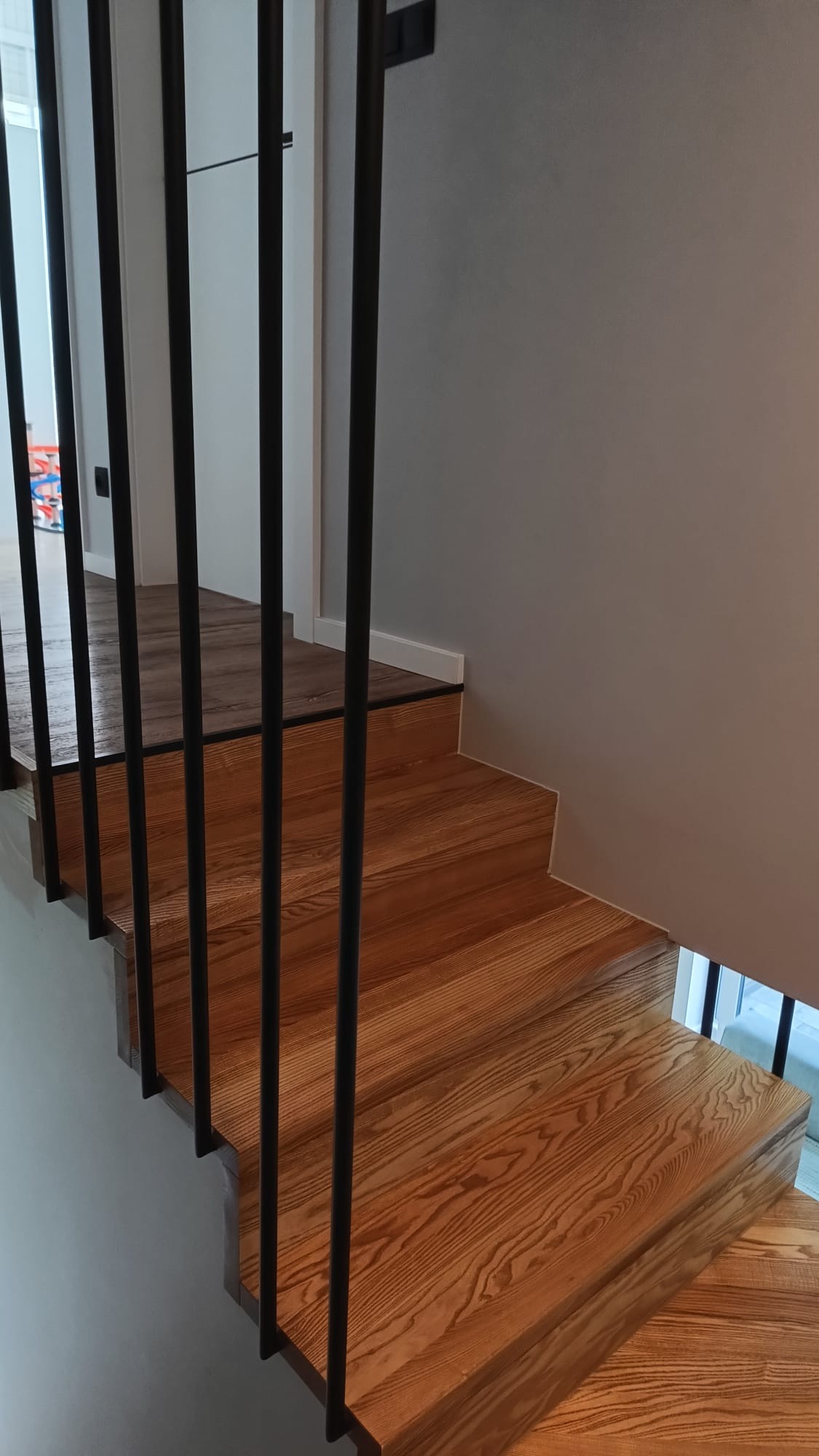 Drewniane schody z czarną balustradą, widok z dołu. Naturalne oświetlenie podkreśla strukturę drewna. Minimalistyczny design, szare ściany w tle.