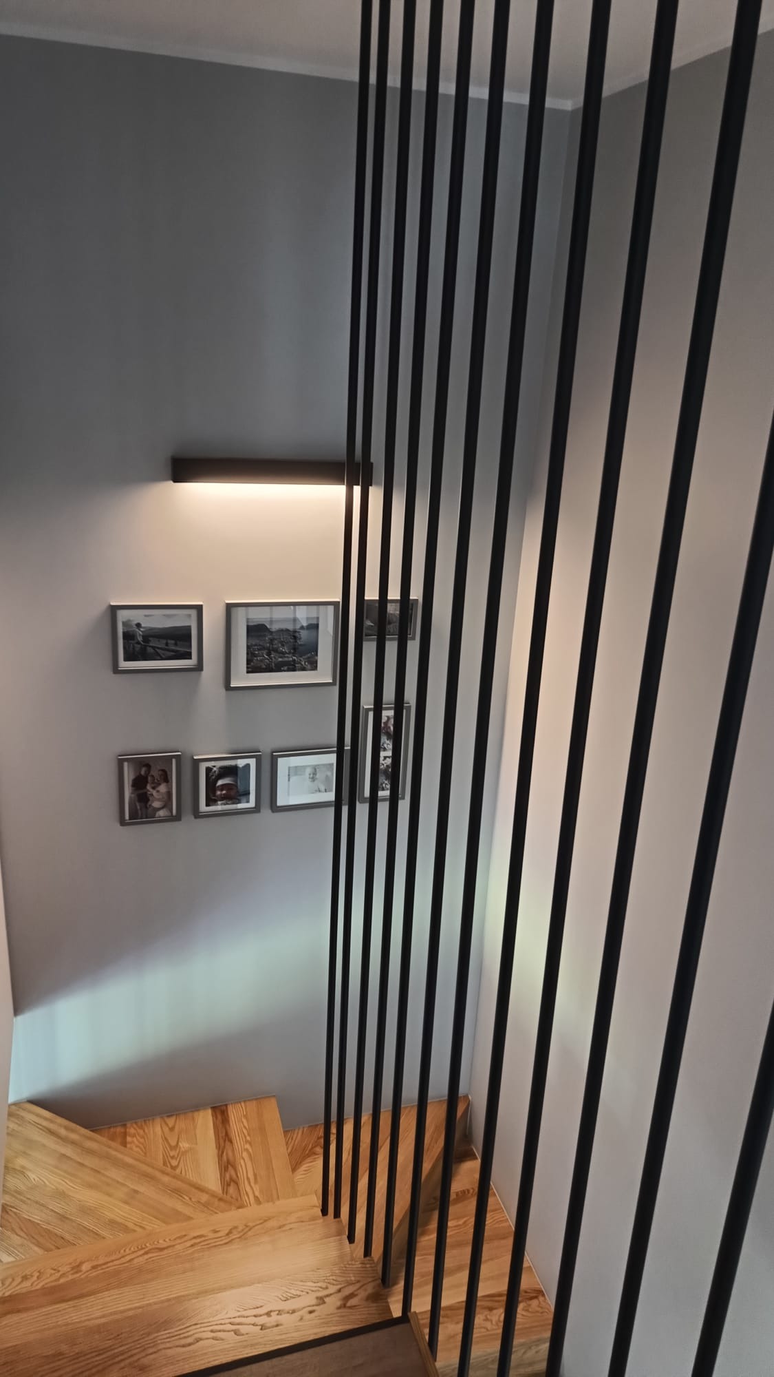 Nowoczesne schody drewniane z czarną, minimalistyczną balustradą. Na ścianie galeria zdjęć podświetlona listwą LED. Jasne drewno stopni kontrastuje z szarością ściany.