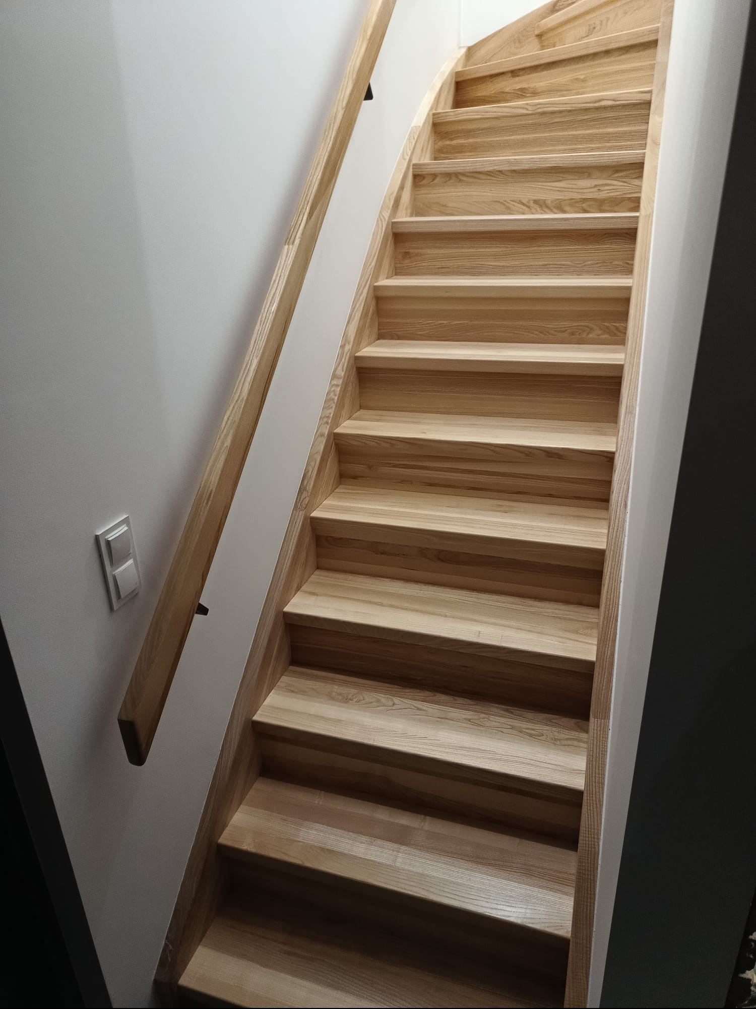 Jasne, drewniane schody z zakrętem i prostą, drewnianą barierką, zamontowane w jasnym wnętrzu. Widoczny włącznik światła na ścianie.