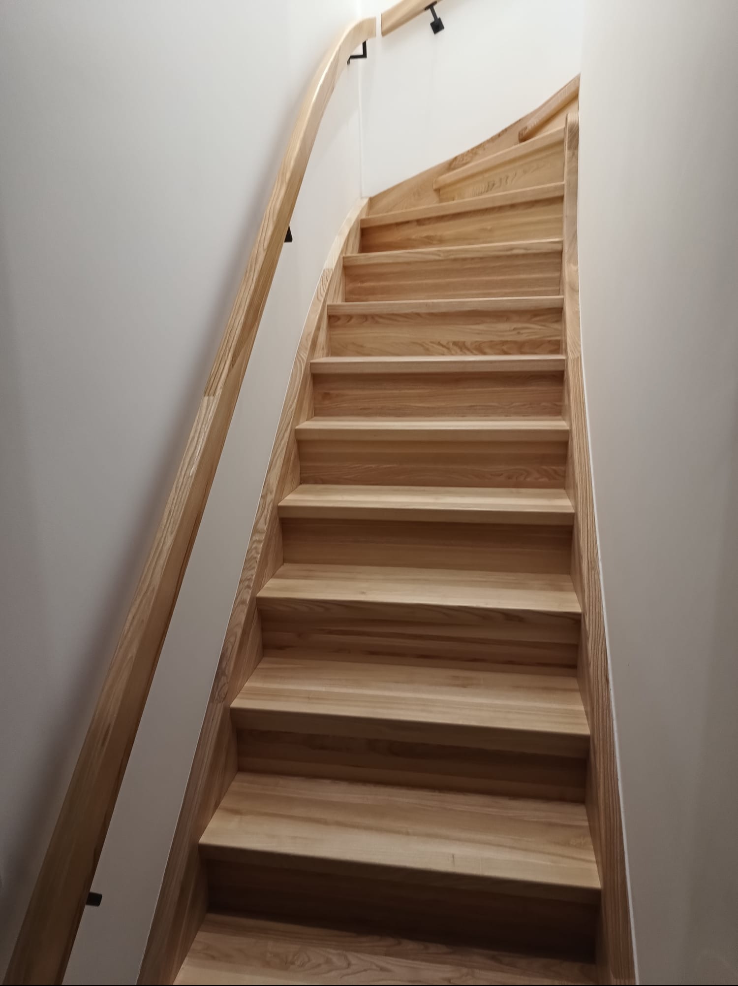 Jasne, drewniane schody z zakrętem i minimalistyczną balustradą, widziane z dołu, oświetlone punktowymi lampami na suficie. Nowoczesny design.