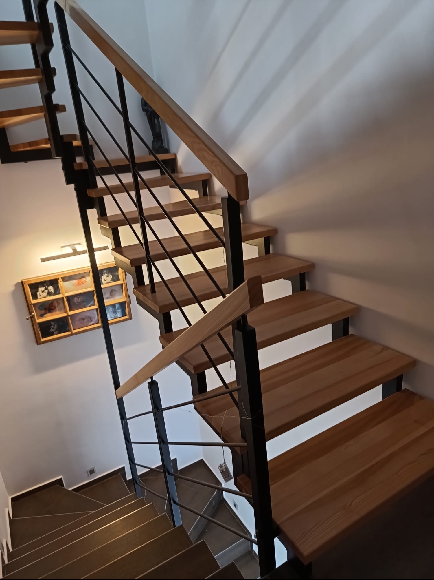 Nowoczesne schody drewniane z czarnymi metalowymi barierkami, oświetlone ciepłym światłem, z widoczną galerią zdjęć na ścianie. Ujęcie z dołu, podkreślające konstrukcję.