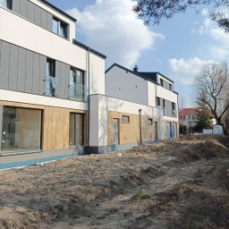 AD-BUD usługi remontowo budowlane - Nowoczesny budynek w trakcie wykończenia elewacji: białe ściany, szare panele, drewniane elementy. Widoczne przygotowane podłoże i fragmenty instalacji.