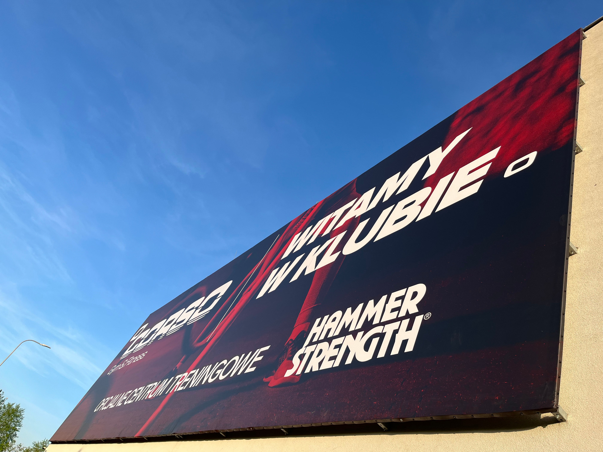 Billboard reklamowy klubu fitness z hasłem 'Witamy w klubie' i logotypami 'Corseo' i 'Hammer Strength' na tle błękitnego nieba, widziany z dołu pod kątem.