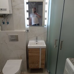 Domex - Nowoczesna łazienka z kabiną prysznicową, umywalką z szafką w odcieniach drewna i podwieszaną toaletą. W lustrze odbija się osoba robiąca zdjęcie.