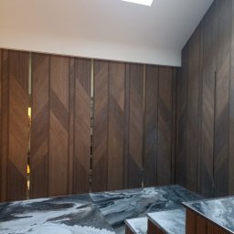 Domex - Nowoczesne wykończenie wnętrza z drewnianą ścianą o geometrycznym wzorze i marmurowym blatem. Widoczne oświetlenie sufitowe.