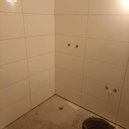 Domex - Wykończona ściana w łazience z białymi, kwadratowymi płytkami. Widoczne przygotowane przyłącza wodne i wiadro z zaprawą. Podłoga surowa, przed wykończeniem.