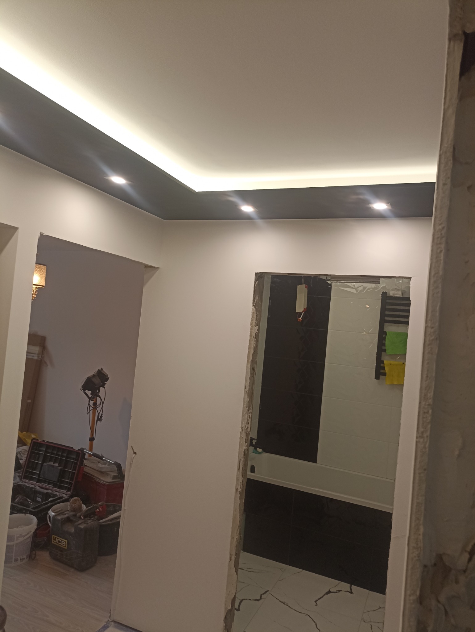 Remont łazienki w trakcie: widok na wykończoną, czarno-białą łazienkę przez surowy otwór drzwiowy. Sufit podwieszany z oświetleniem LED. W tle narzędzia i sprzęt remontowy.