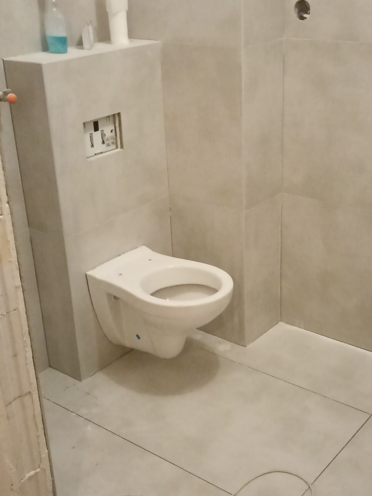 Zamontowana podwieszana toaleta w łazience wykończonej jasnymi, kwadratowymi płytkami. Widoczna instalacja podtynkowa i fragment ściany z niedokończonym wykończeniem.