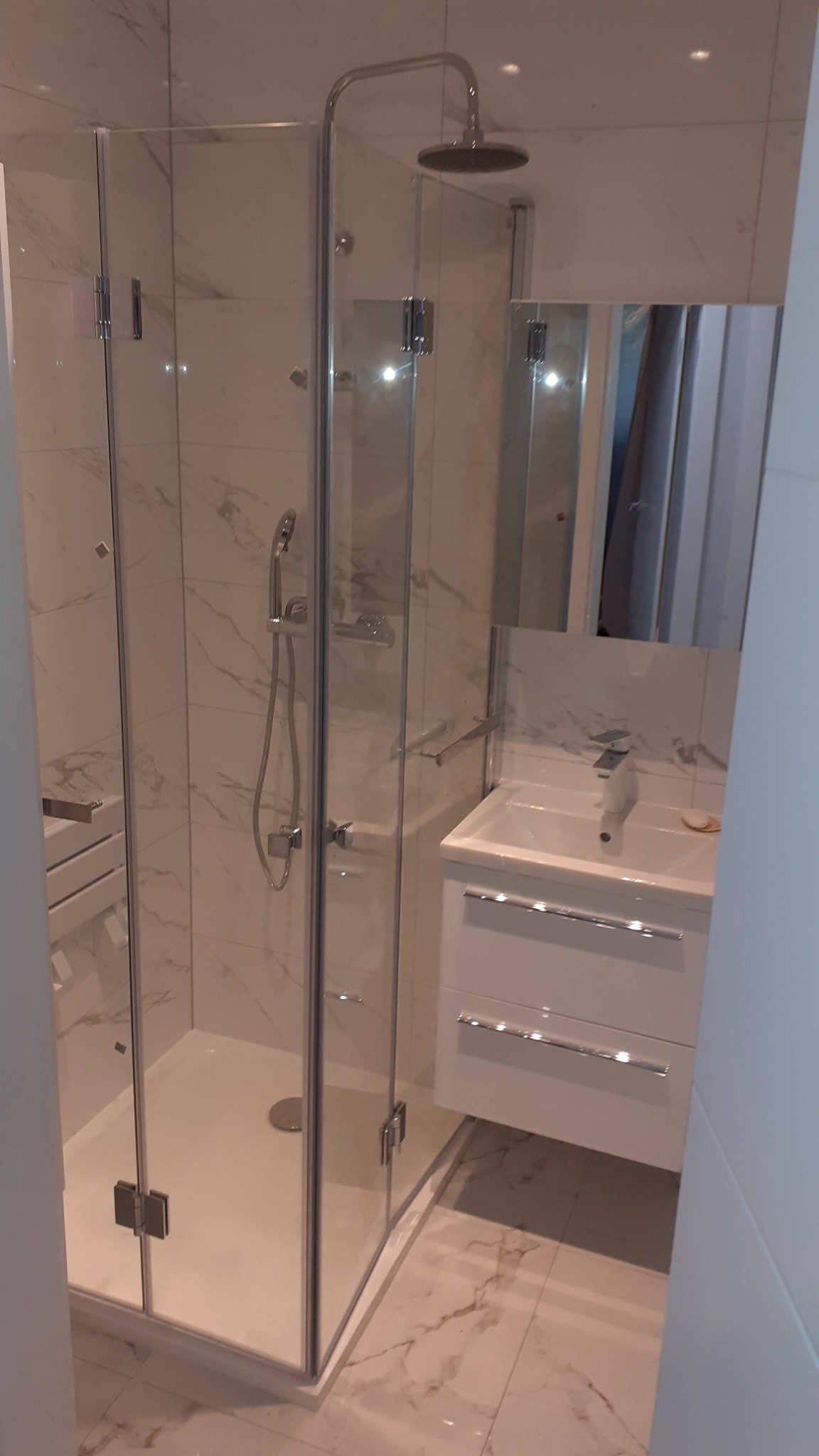 Nowoczesna kabina prysznicowa z deszczownicą, biała umywalka z szafką i marmurowe płytki w wykończonej łazience. Minimalistyczny design, chromowane detale.