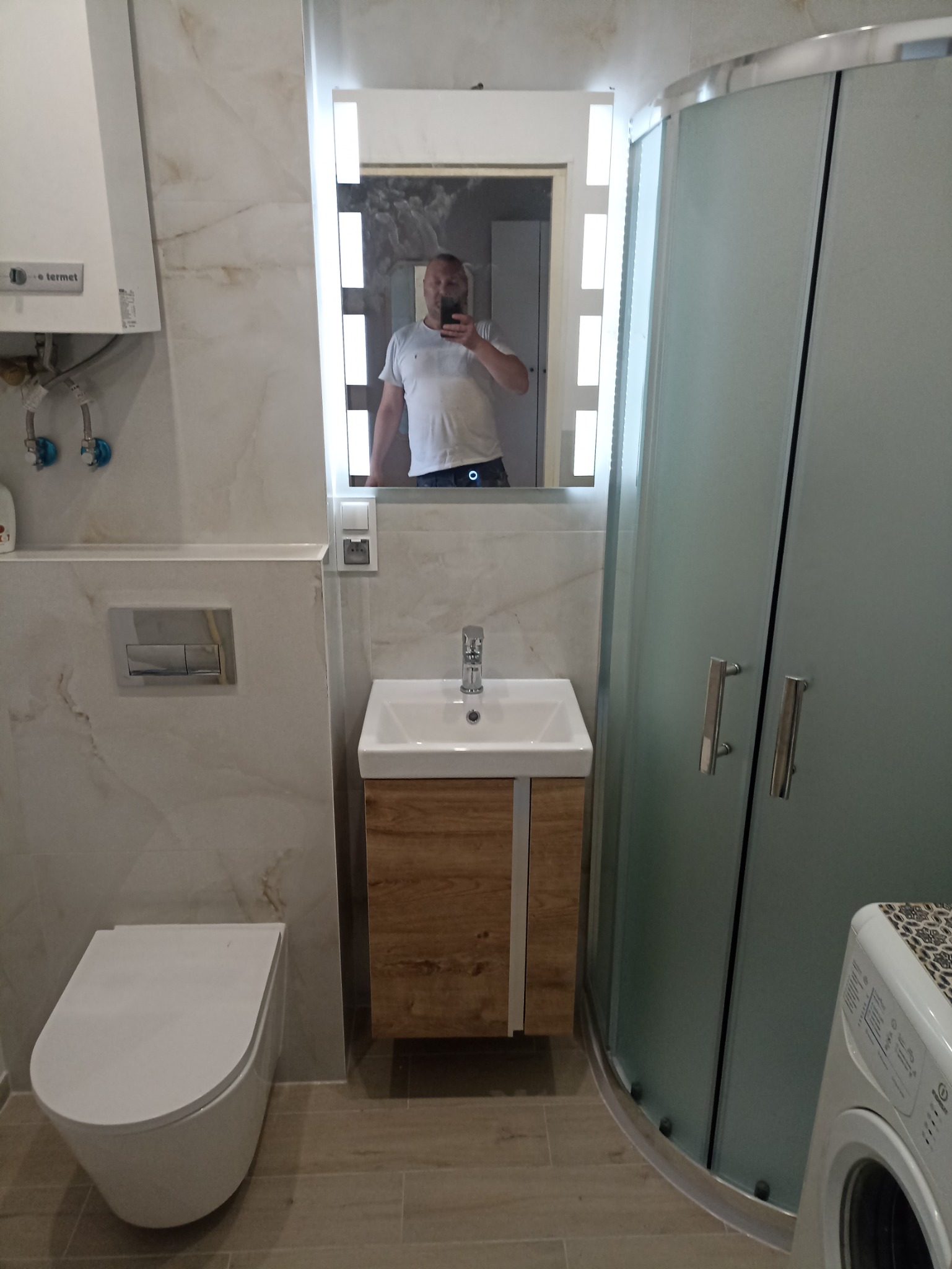 Nowoczesna łazienka z kabiną prysznicową, umywalką z szafką w odcieniach drewna i podwieszaną toaletą. W lustrze odbija się osoba robiąca zdjęcie.