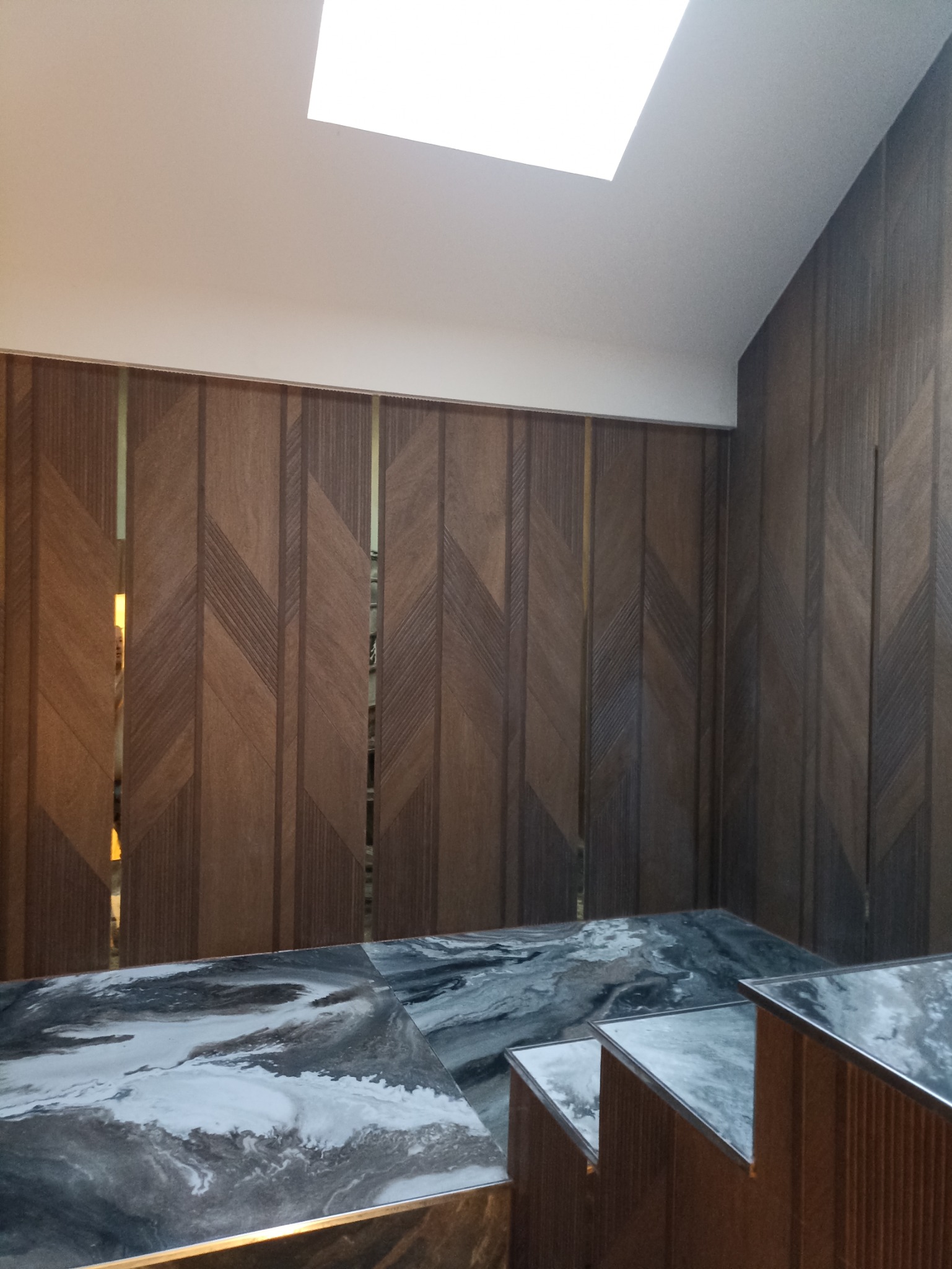 Nowoczesne wykończenie wnętrza z drewnianą ścianą o geometrycznym wzorze i marmurowym blatem. Widoczne oświetlenie sufitowe.