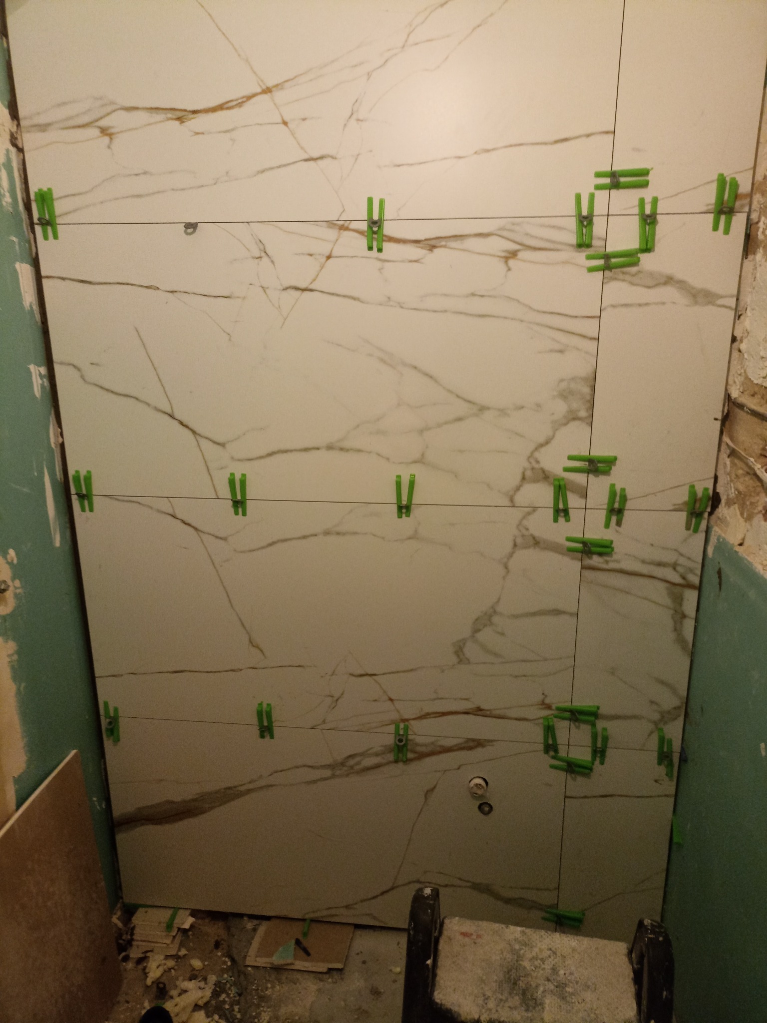 Ściana z płytek imitujących marmur, z zielonymi klipsami poziomującymi i sznurkiem, w trakcie układania. Widoczne surowe ściany i podłoga w trakcie prac.