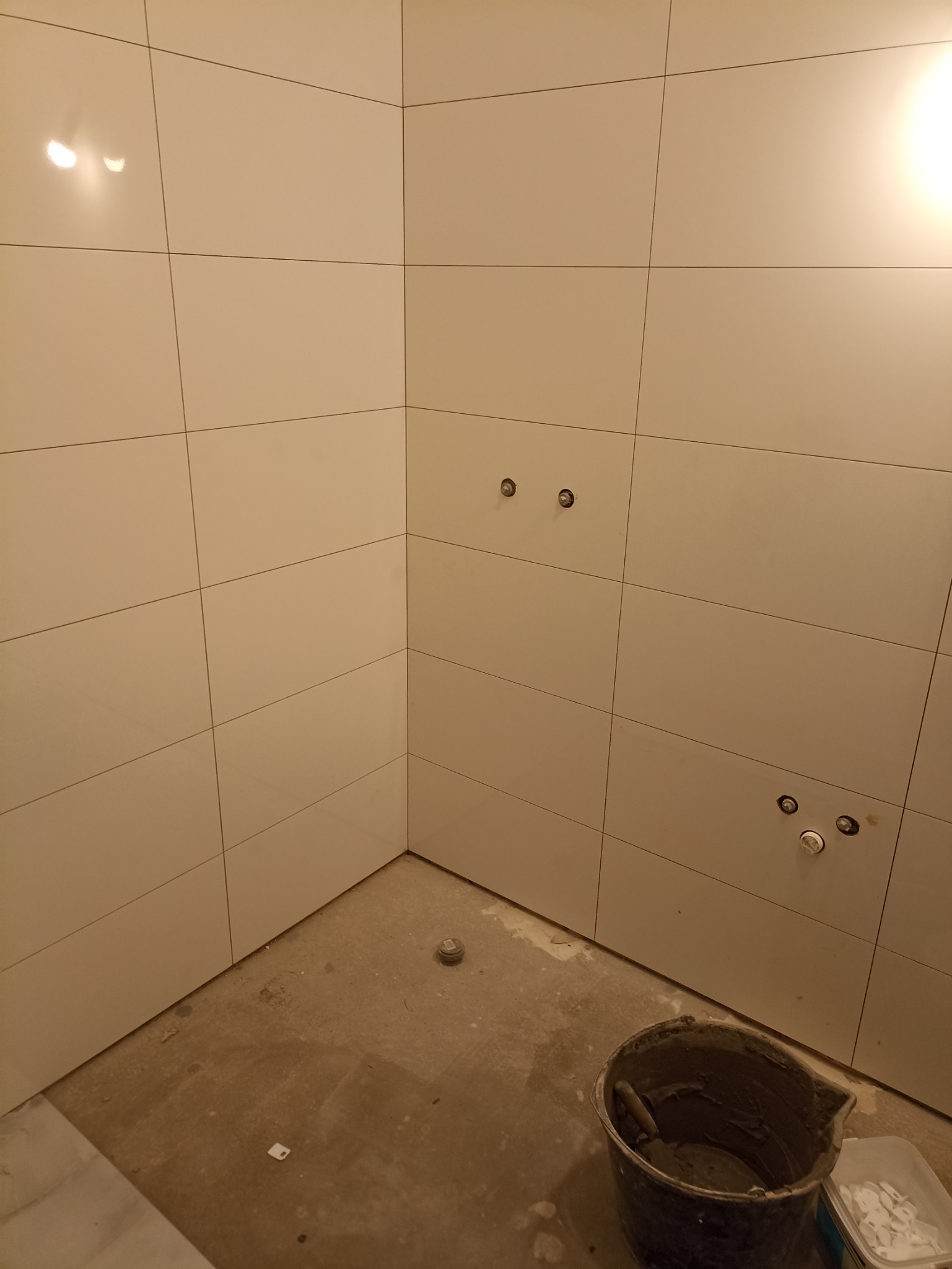 Wykończona ściana w łazience z białymi, kwadratowymi płytkami. Widoczne przygotowane przyłącza wodne i wiadro z zaprawą. Podłoga surowa, przed wykończeniem.