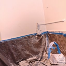 Szymon Wasilewski - Przygotowanie do malowania: ściana zabezpieczona folią i taśmą malarską, rura osłonięta. Widoczne ślady farby. Ochrona podłoża przed zabrudzeniem.