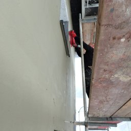 Szymon Wasilewski - Praca na rusztowaniu: aplikacja gładzi na elewacji budynku z użyciem pacy. Widoczne fragmenty okna i ocieplenia. Ujęcie z dołu, podkreślające wysokość ściany.