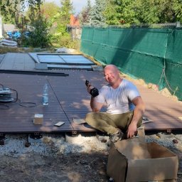 Szymon Wasilewski - Mężczyzna z wkrętarką siedzi na częściowo ułożonym tarasie z desek kompozytowych na zewnątrz. Widoczne podpory tarasu, narzędzia i butelka wody.