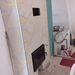 Obudowa kominka z płyt marmurowych w trakcie montażu, zabezpieczona taśmą. Widoczne otwory na wkład kominkowy i wentylację, fragment ściany w kolorze turkusowym.