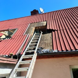 Szymon Wasilewski - Częściowo zdjęty, czerwony dach z eternitu, drabina oparta o ścianę, widoczne okno dachowe i komin z anteną satelitarną na tle błękitnego nieba.