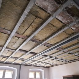 Szymon Wasilewski - Sufit podwieszany w trakcie montażu: metalowa konstrukcja, izolacja z wełny mineralnej, widoczne elementy starego stropu. Ujęcie z dołu, szeroki kąt.