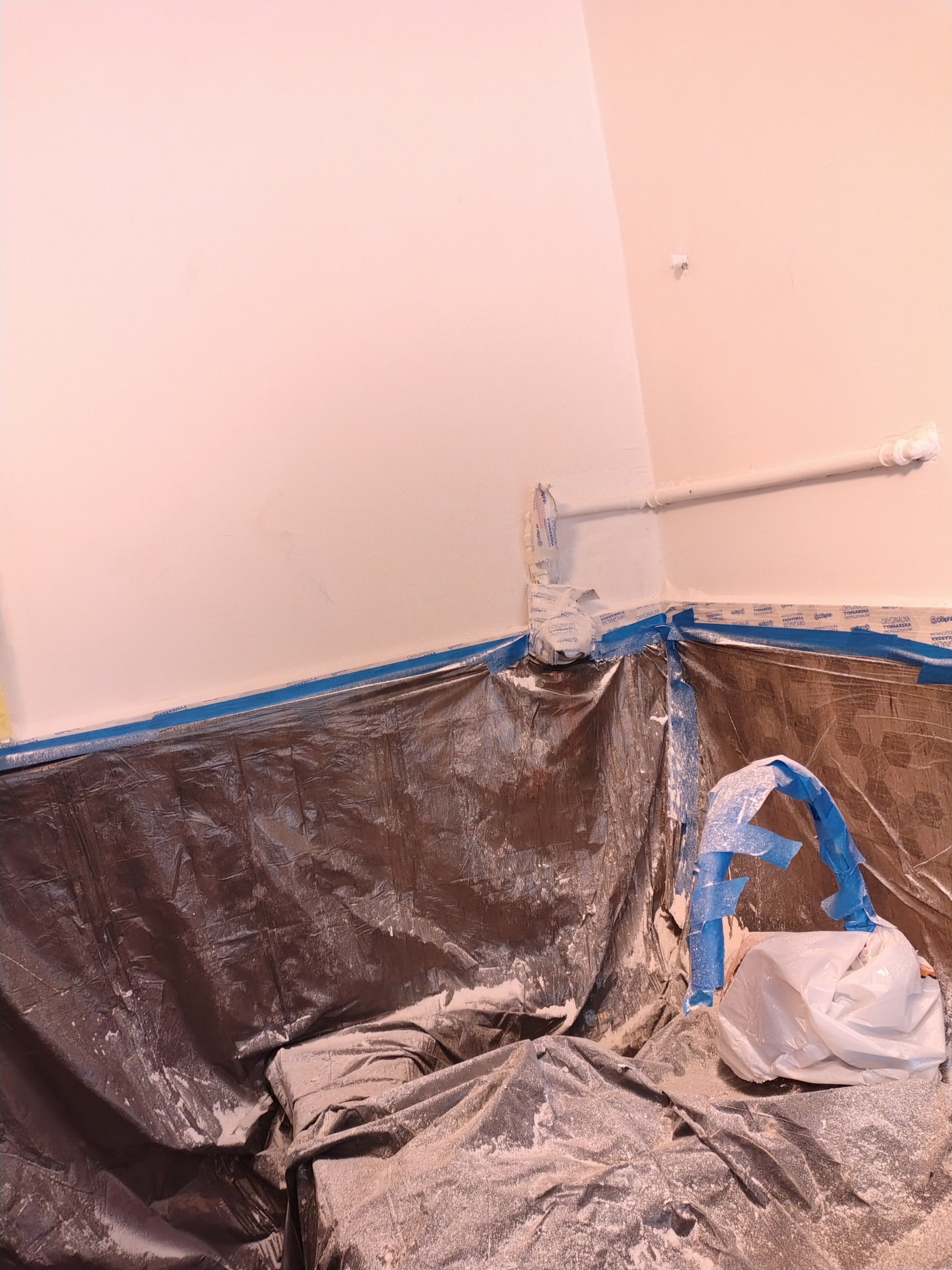 Przygotowanie do malowania: ściana zabezpieczona folią i taśmą malarską, rura osłonięta. Widoczne ślady farby. Ochrona podłoża przed zabrudzeniem.