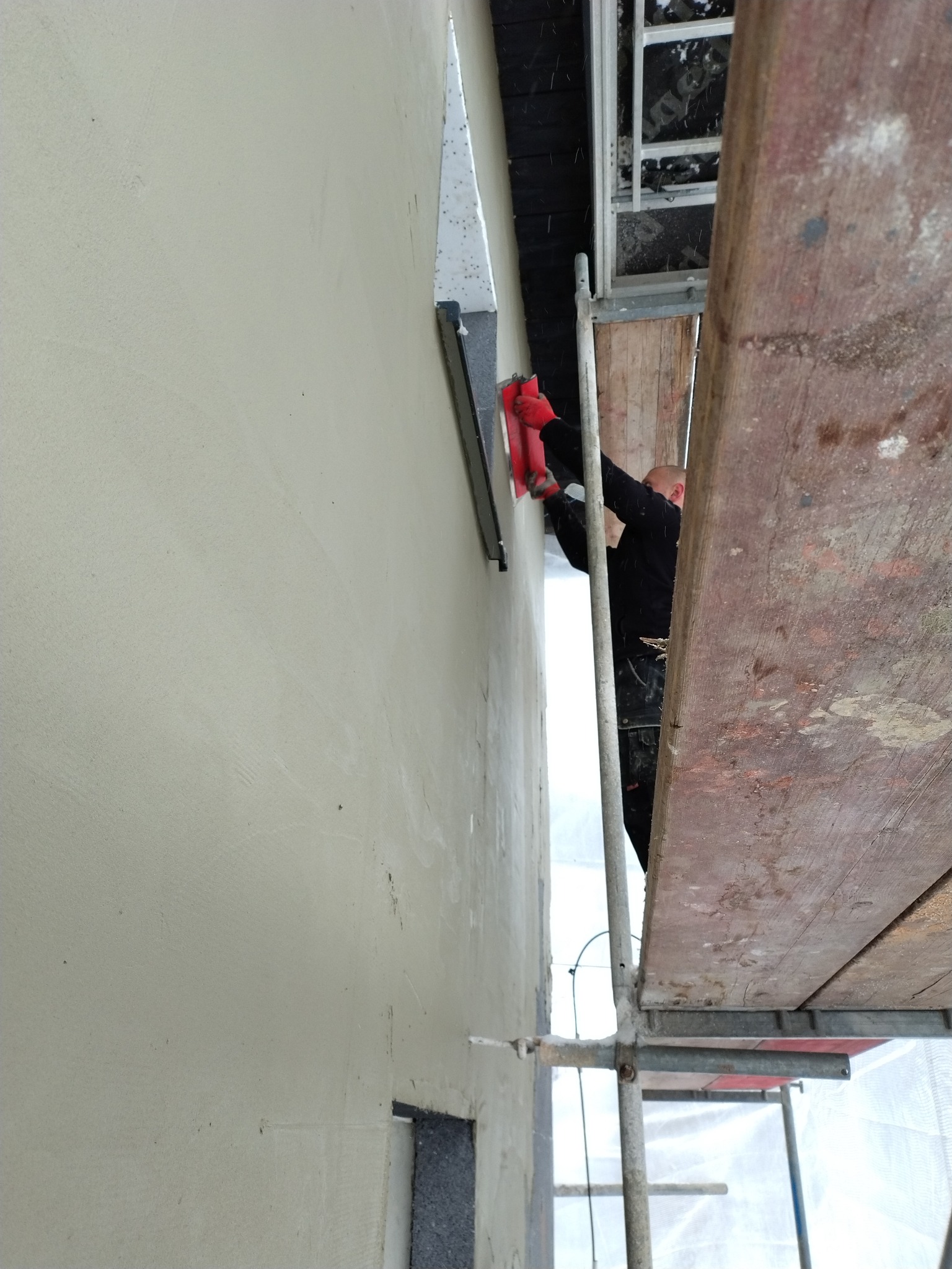 Praca na rusztowaniu: aplikacja gładzi na elewacji budynku z użyciem pacy. Widoczne fragmenty okna i ocieplenia. Ujęcie z dołu, podkreślające wysokość ściany.