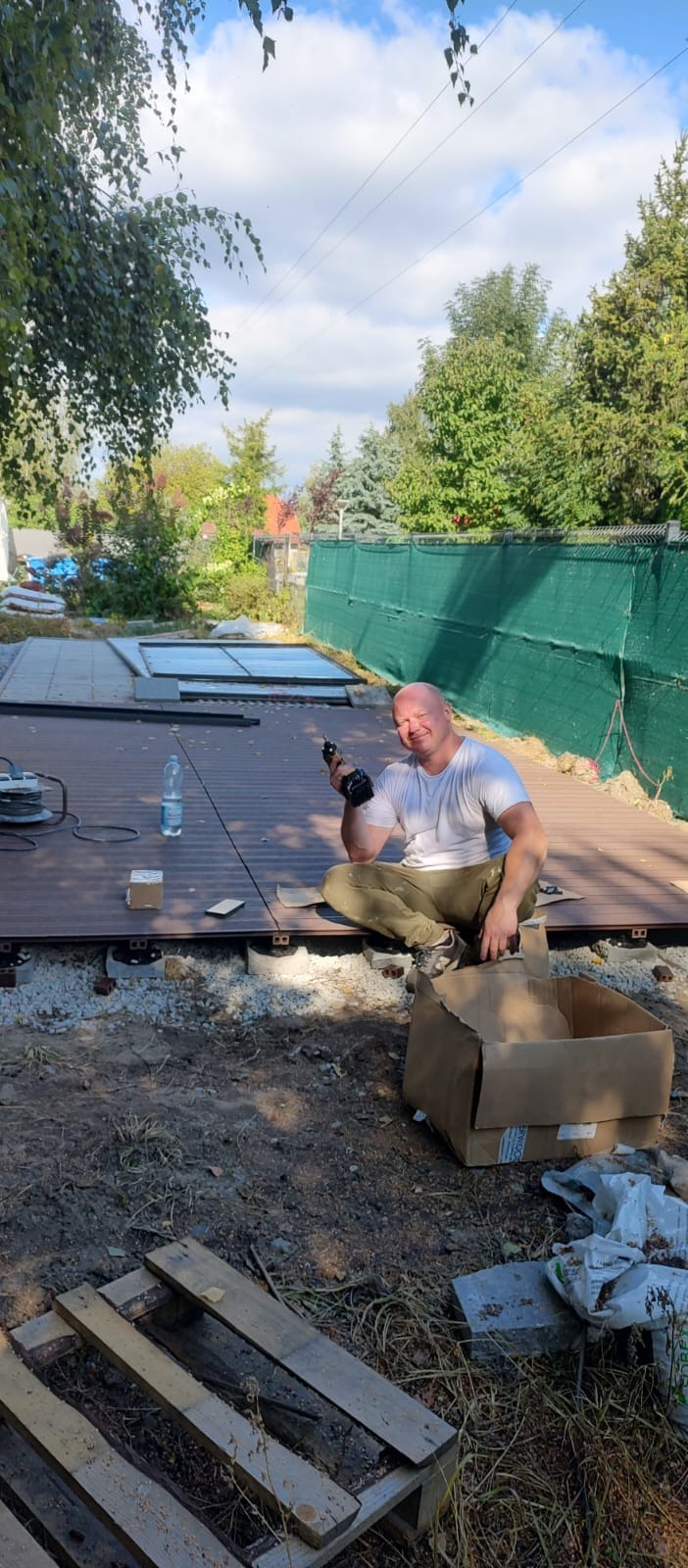 Mężczyzna z wkrętarką siedzi na częściowo ułożonym tarasie z desek kompozytowych na zewnątrz. Widoczne podpory tarasu, narzędzia i butelka wody.