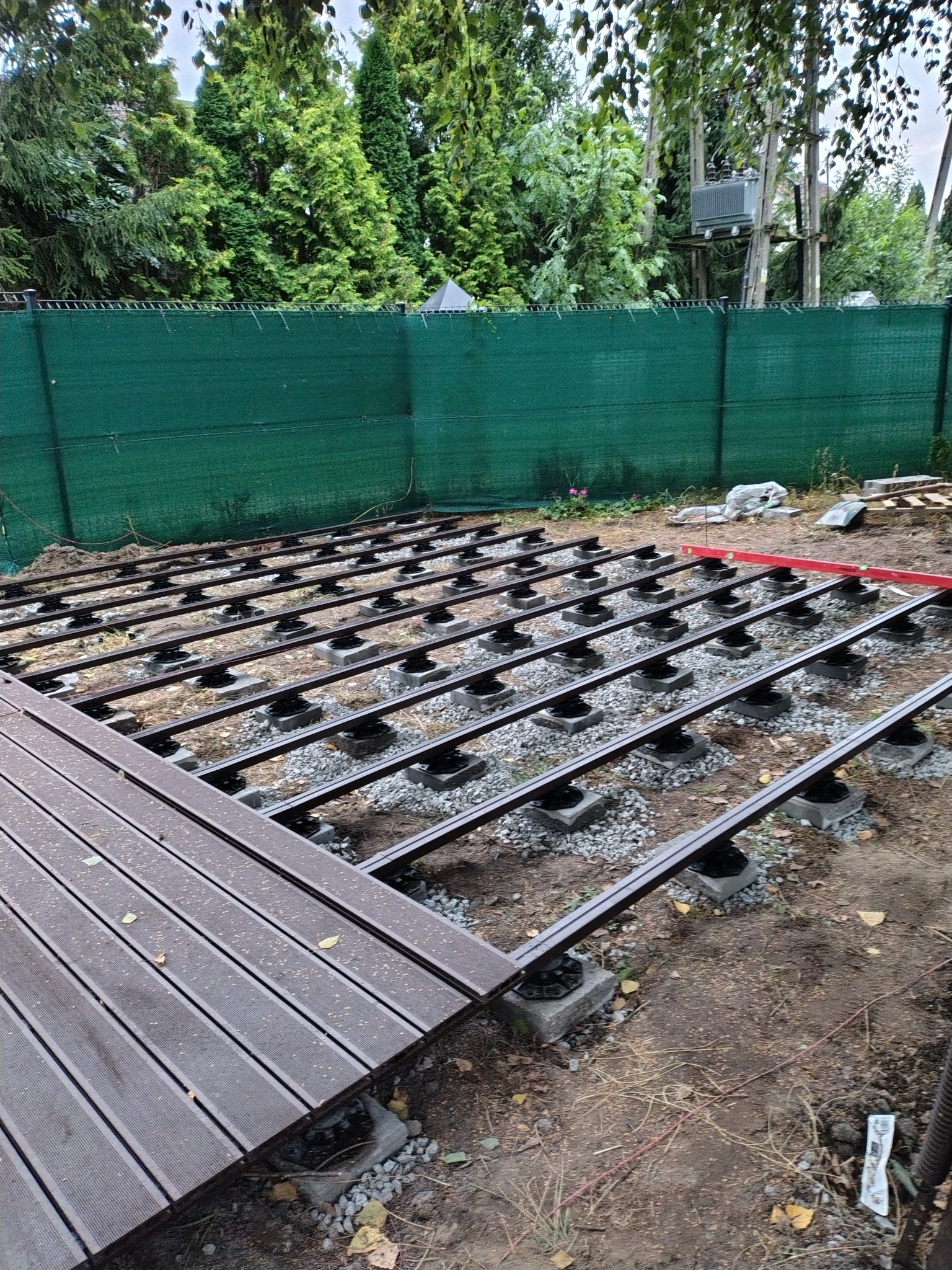 Konstrukcja tarasu kompozytowego w trakcie montażu: legary na regulowanych wspornikach, częściowo ułożone deski. Widoczny poziomica i zielone ogrodzenie w tle.