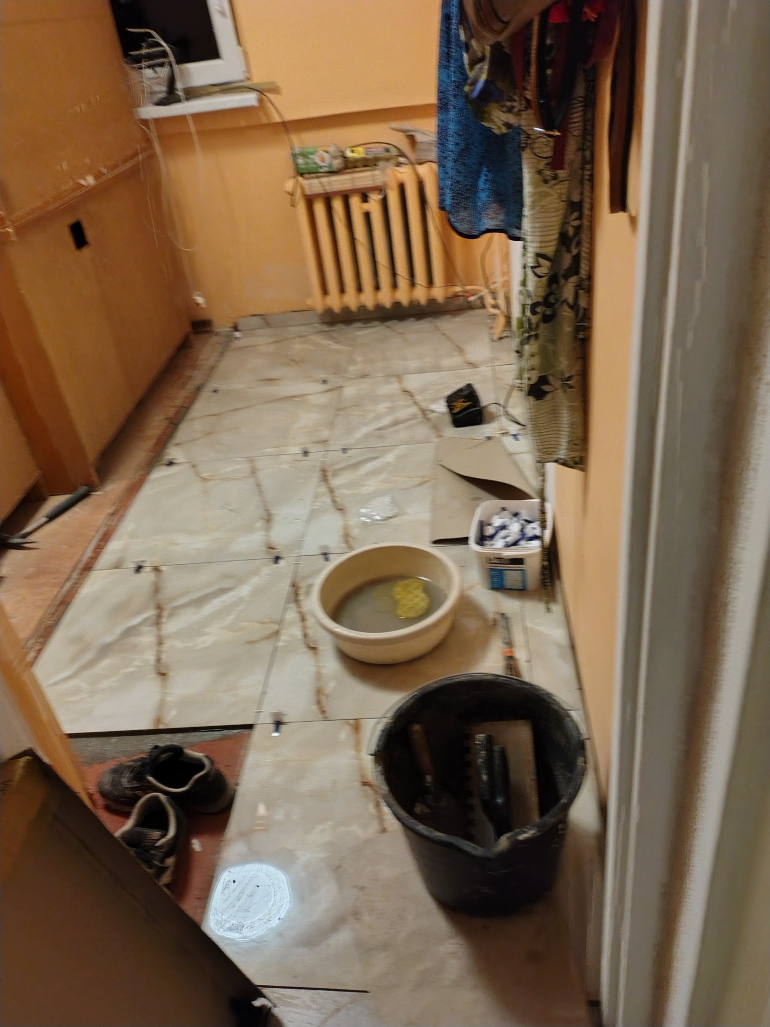 Układanie płytek podłogowych w trakcie prac. Widoczne narzędzia, wiadro z wodą, krzyżyki dystansowe i częściowo ułożone płytki w beżowym odcieniu.