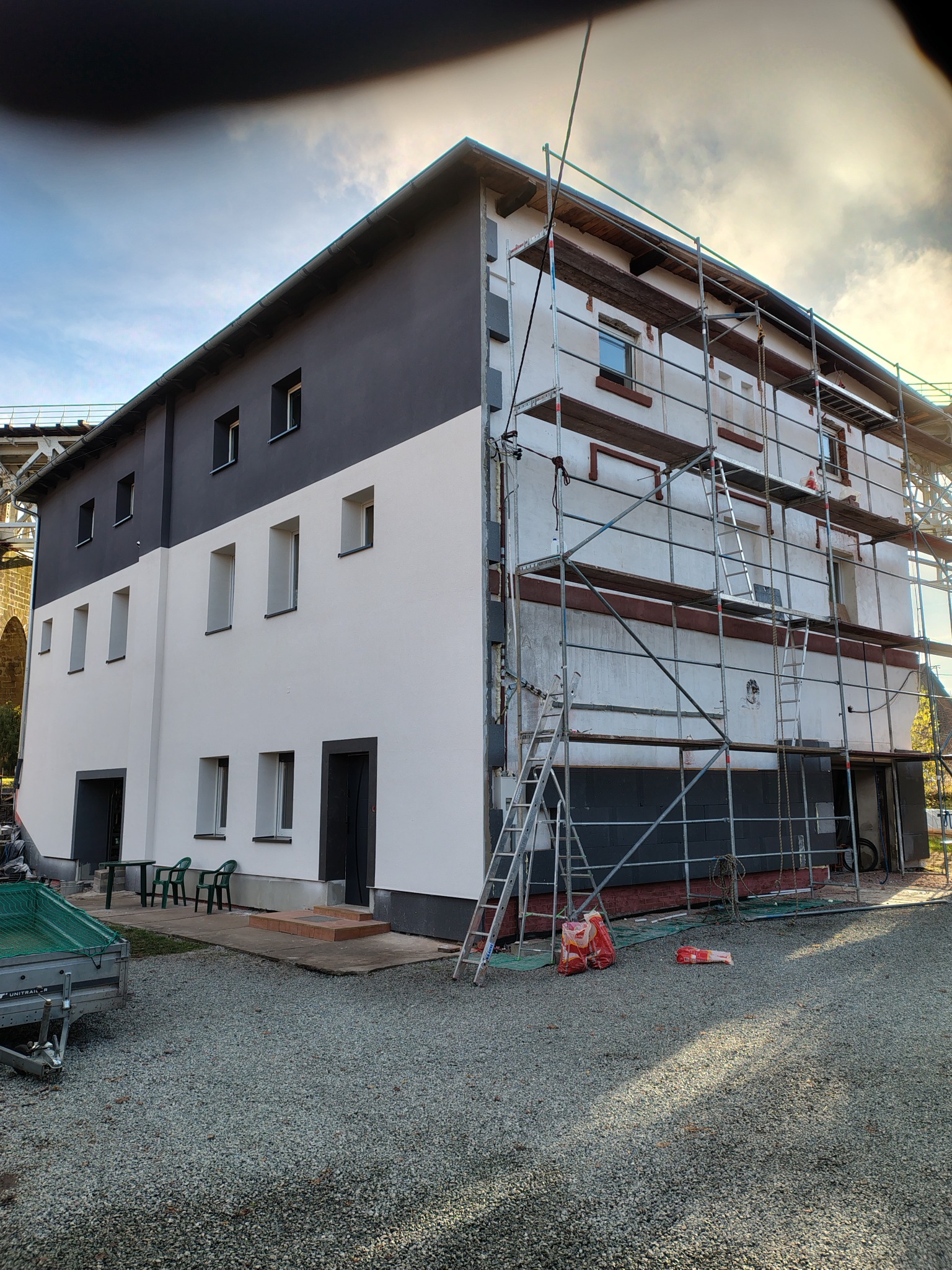 Budynek po częściowym malowaniu elewacji na szaro i biało, z rusztowaniem z boku. Widoczne worki z zaprawą i przyczepka. W tle konstrukcja mostu.