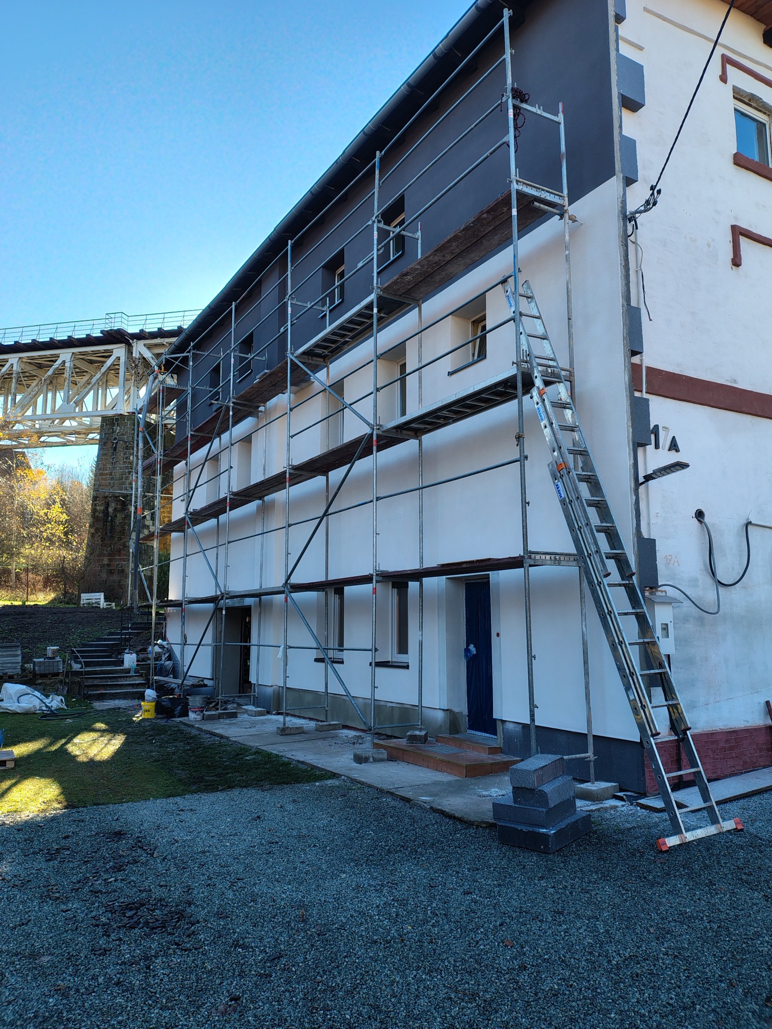 Elewacja budynku w trakcie malowania, widoczne rusztowania i drabina. Część budynku pomalowana na szaro, druga na biało. W tle widoczny most.