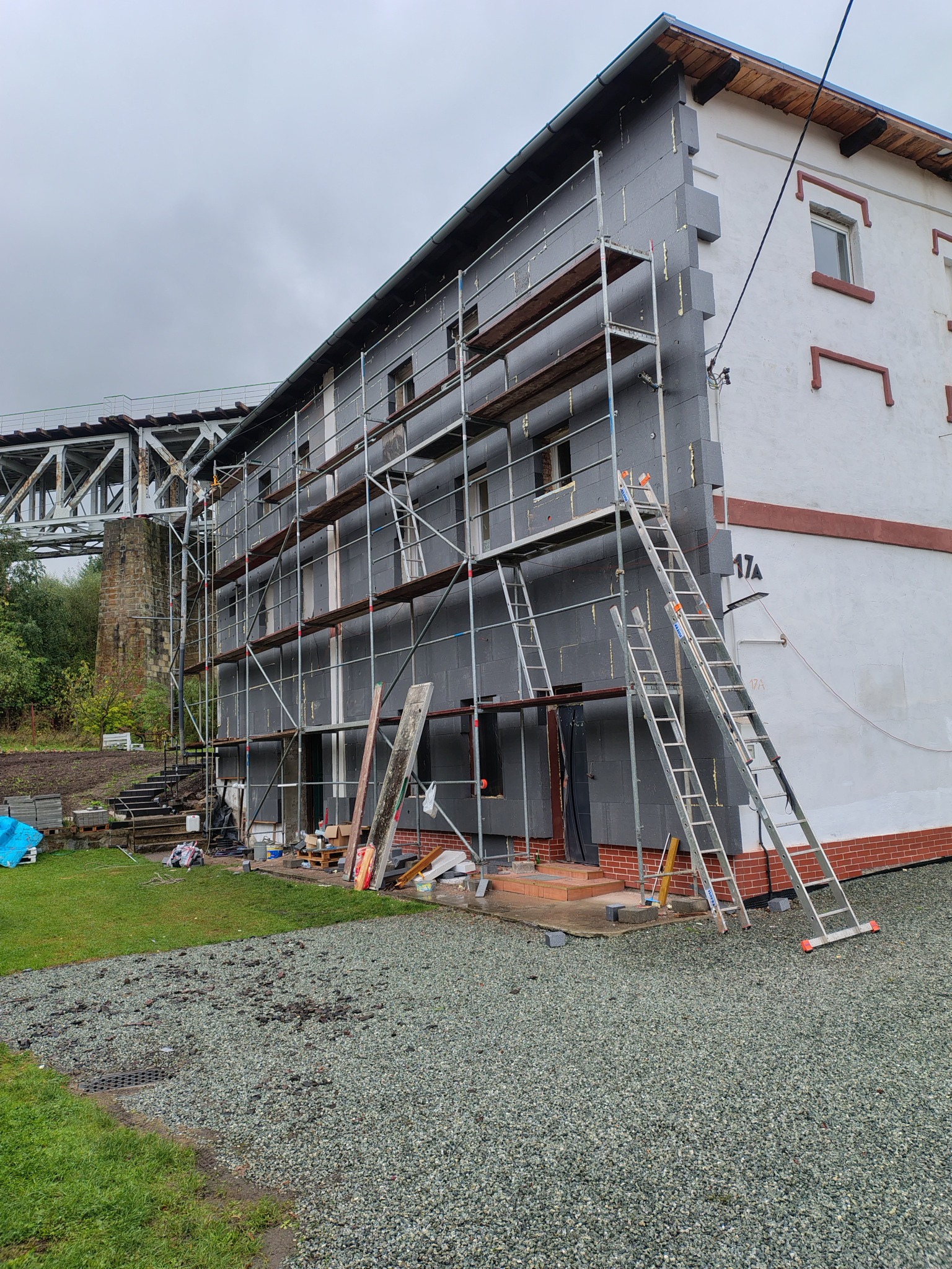 Budynek w trakcie ocieplania styropianem, widoczne rusztowania i drabiny. W tle konstrukcja mostu. Prace elewacyjne w toku, szary styropian na ścianie.