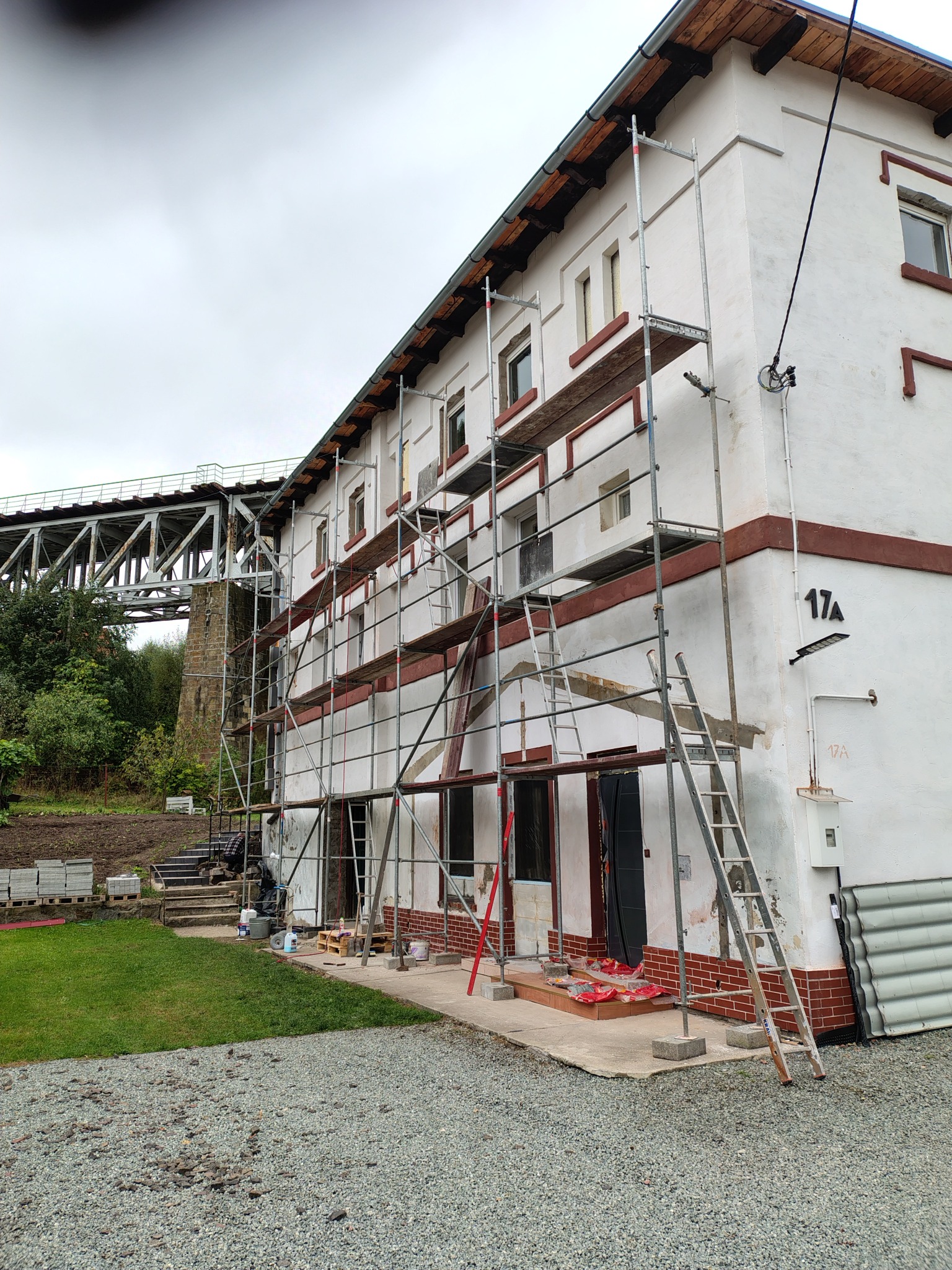 Budynek z rusztowaniem w trakcie prac elewacyjnych, widoczny fragment mostu w tle. Numer budynku 17A. Prace przygotowawcze do malowania.