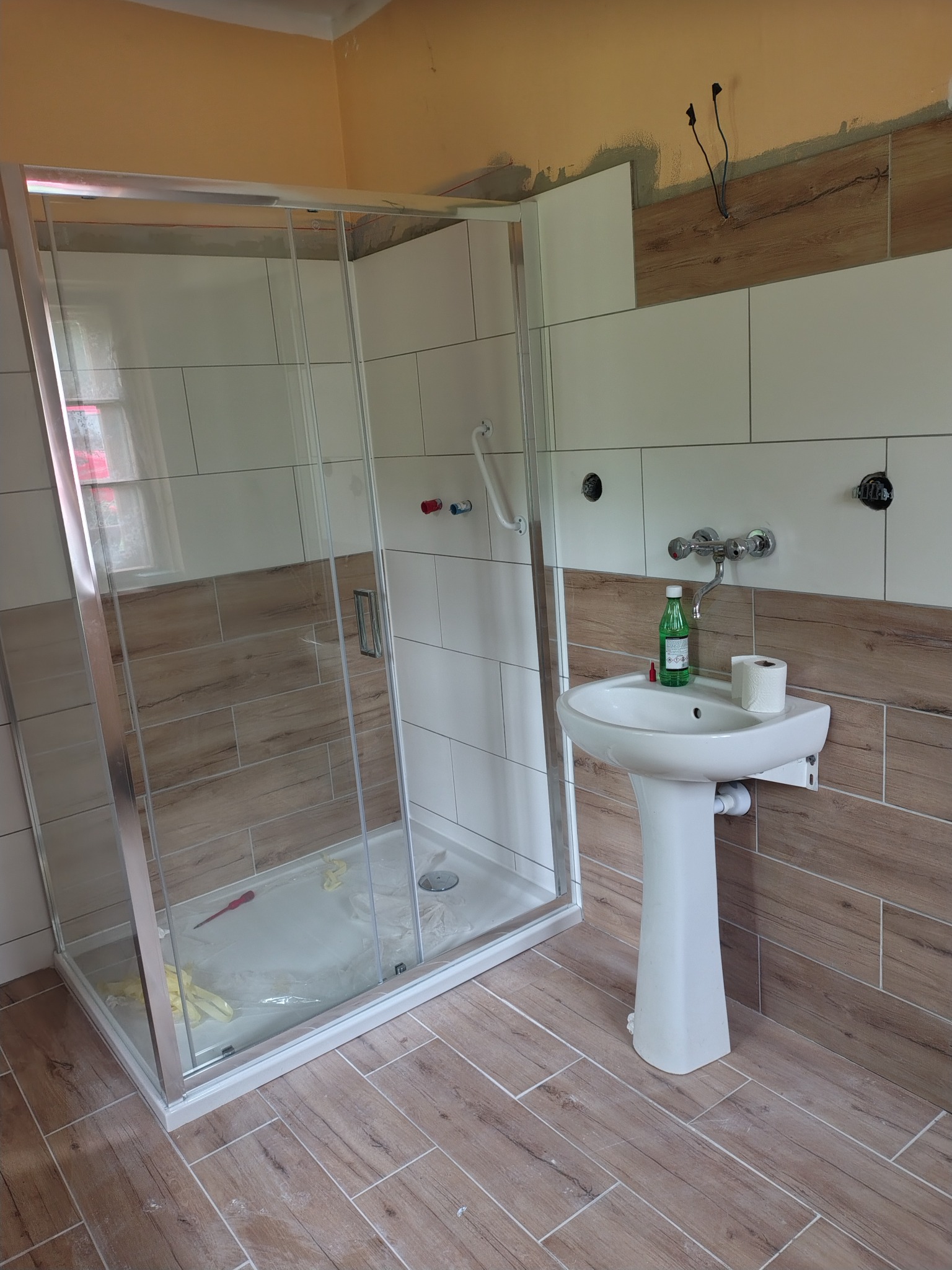 Wykończona łazienka z kabiną prysznicową, umywalką na postumencie i płytkami imitującymi drewno na podłodze i ścianach. Widoczne niedokończone prace na górze ściany.