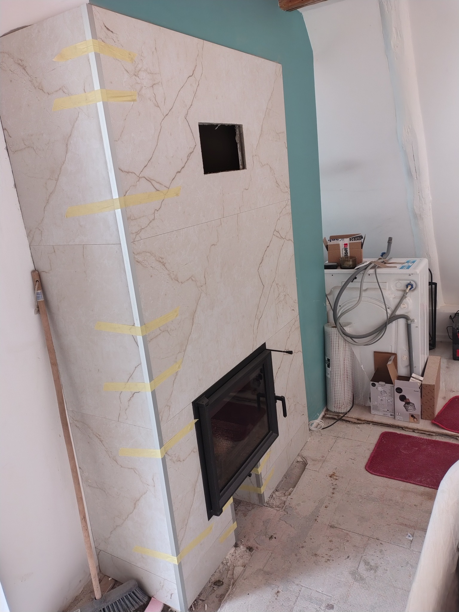 Obudowa kominka z płyt marmurowych w trakcie montażu, zabezpieczona taśmą. Widoczne otwory na wkład kominkowy i wentylację, fragment ściany w kolorze turkusowym.