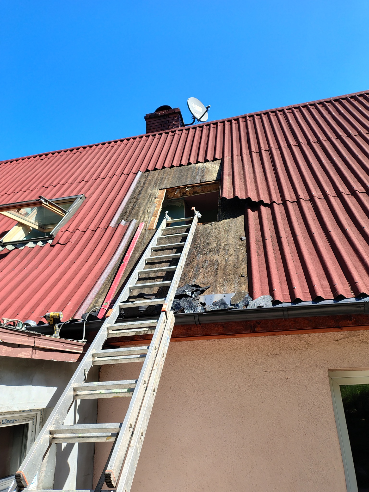 Częściowo zdjęty, czerwony dach z eternitu, drabina oparta o ścianę, widoczne okno dachowe i komin z anteną satelitarną na tle błękitnego nieba.