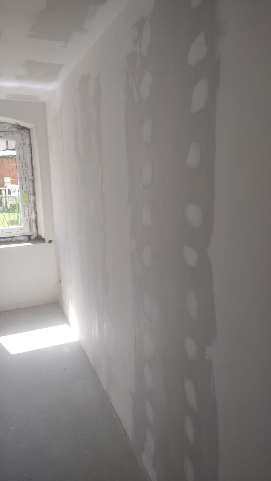 Ściana po szpachlowaniu z widocznymi łatami, przygotowana do dalszych prac wykończeniowych. W rogu okno z widokiem na zewnątrz. Podłoga szara, surowa.