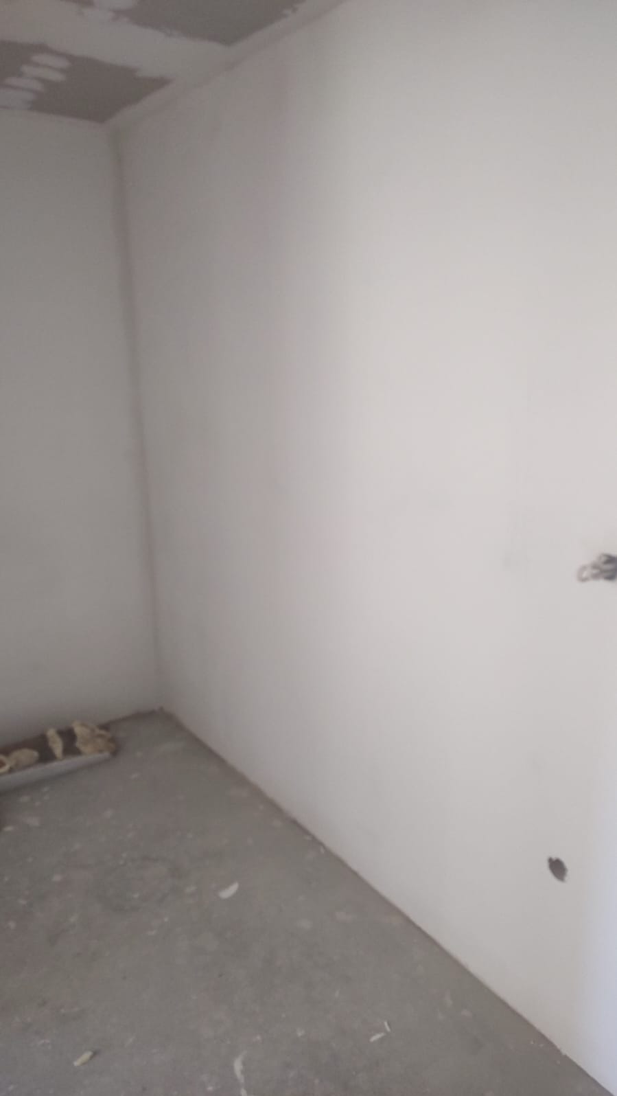 Wnętrze pokoju po szpachlowaniu ścian i sufitu, przygotowane do dalszych prac wykończeniowych. Widoczne gładkie, białe ściany i szary betonowy podkład.