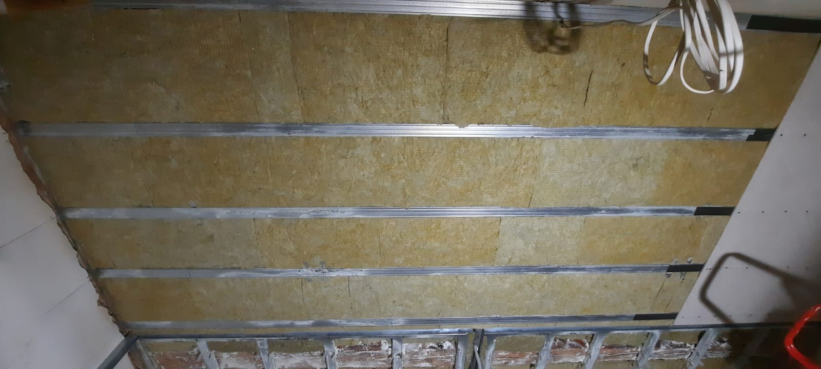 Izolacja poddasza wełną mineralną, widoczne metalowe profile konstrukcyjne. Fragment ściany z płytą gipsową. Prace wykończeniowe na poddaszu.