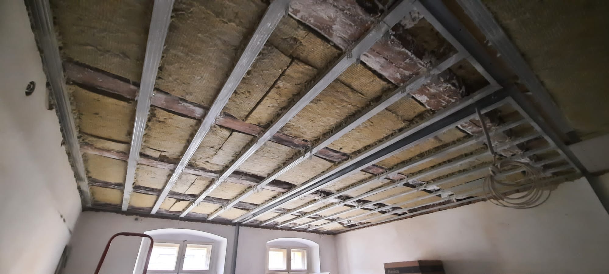 Sufit podwieszany w trakcie montażu: metalowa konstrukcja, izolacja z wełny mineralnej, widoczne elementy starego stropu. Ujęcie z dołu, szeroki kąt.