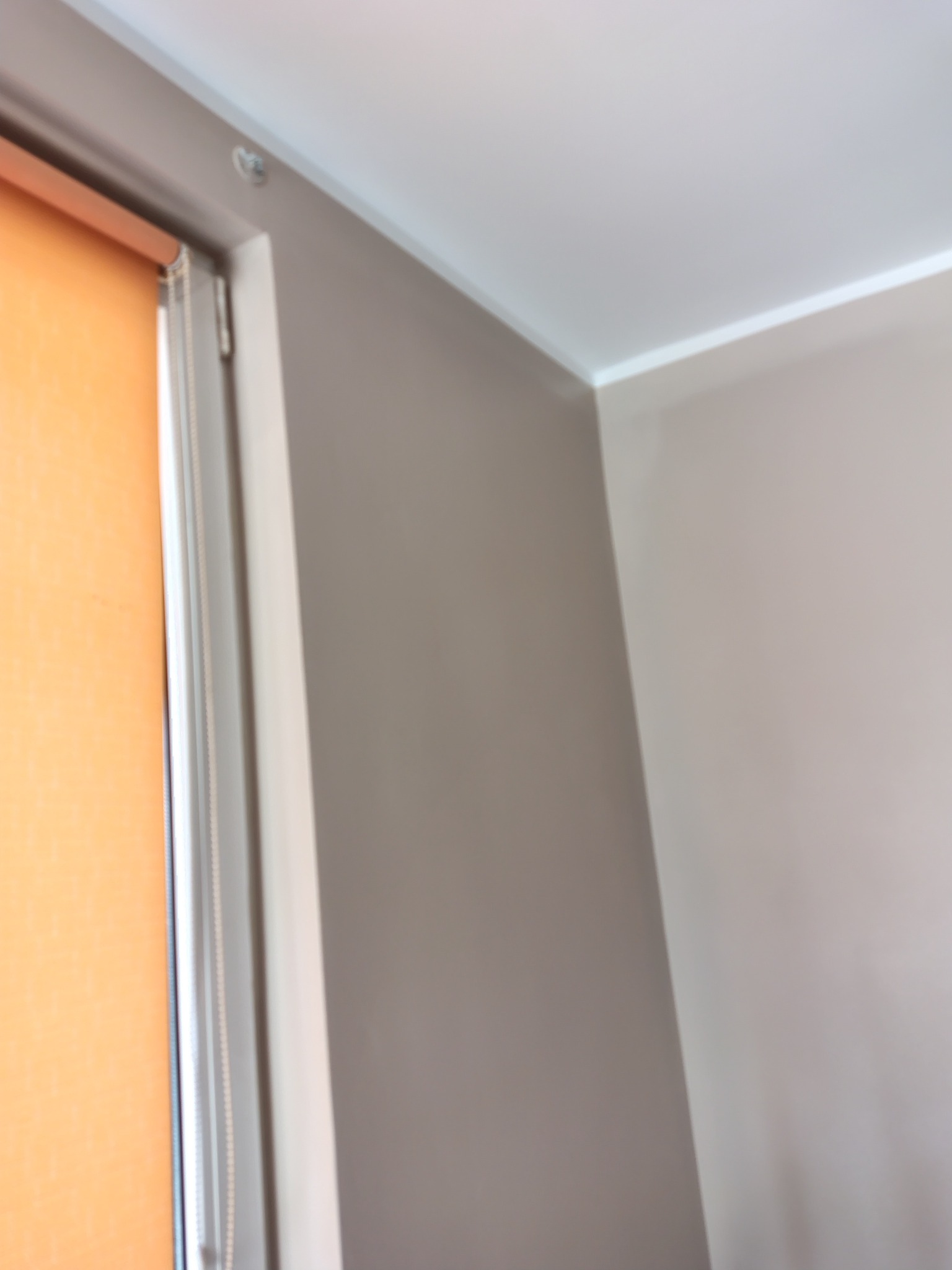 Fragment wnętrza: beżowa ściana po malowaniu, biały sufit z listwą, okno z pomarańczową roletą. Widoczny narożnik pokoju i fragment ramy okiennej.