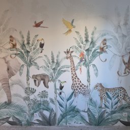 Remonty i wykończenia- Wojciech Rogus - Ściana z fototapetą w motywy dżungli: słoń, żyrafa, małpy, leniwiec i leopard wśród tropikalnych roślin. Widoczne gniazdka elektryczne i fragment surowej ściany.