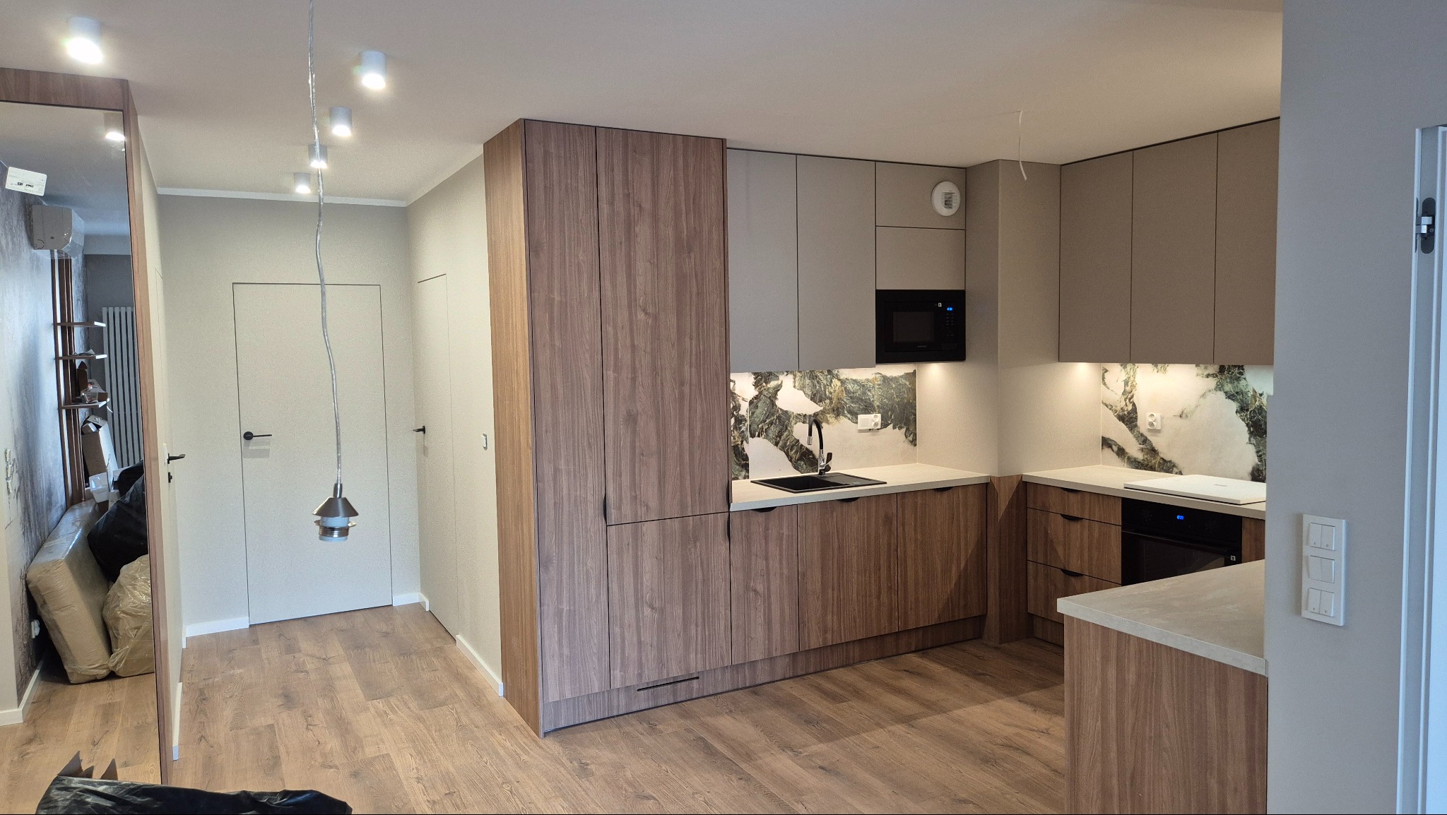 Nowoczesna kuchnia w odcieniach beżu i drewna z marmurowym backsplashiem. Widoczne oświetlenie punktowe i drewniana podłoga. Minimalistyczny design.