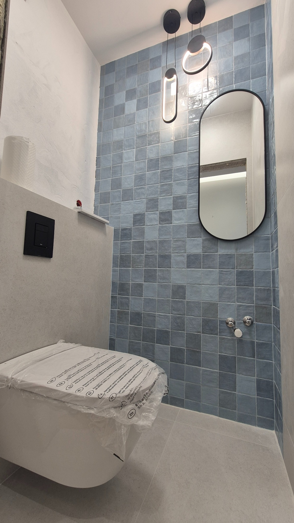 Wykończona łazienka z niebieskimi płytkami, owalnym lustrem i nowoczesnym oświetleniem. Toaleta zabezpieczona folią ochronną. Minimalistyczny design.
