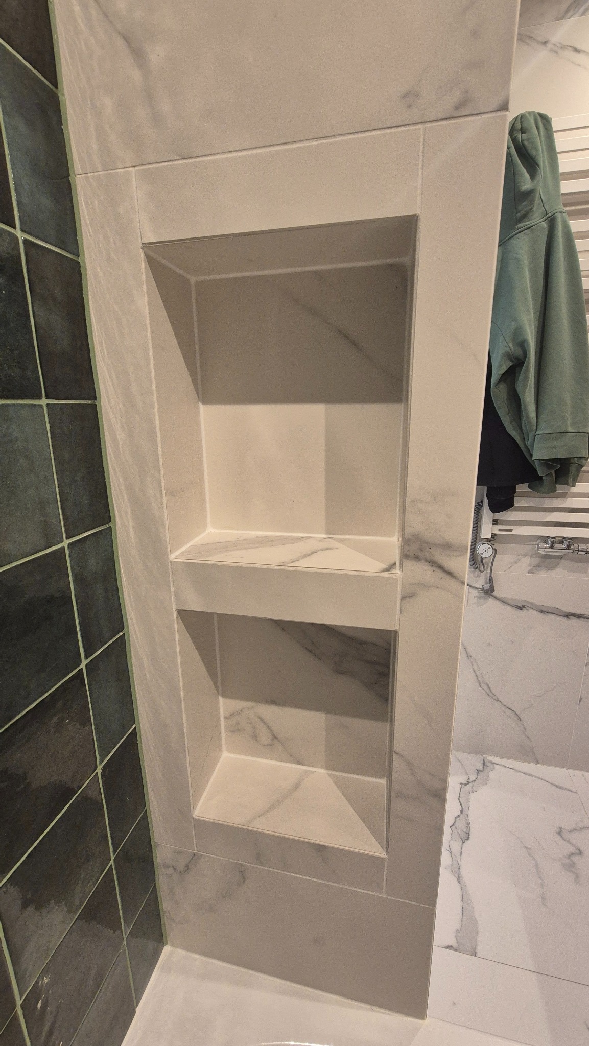 Wnęka z półkami wyłożona płytkami imitującymi marmur, wkomponowana w ścianę łazienki z ciemnymi płytkami po lewej i jasnymi z wzorem marmuru po prawej stronie.