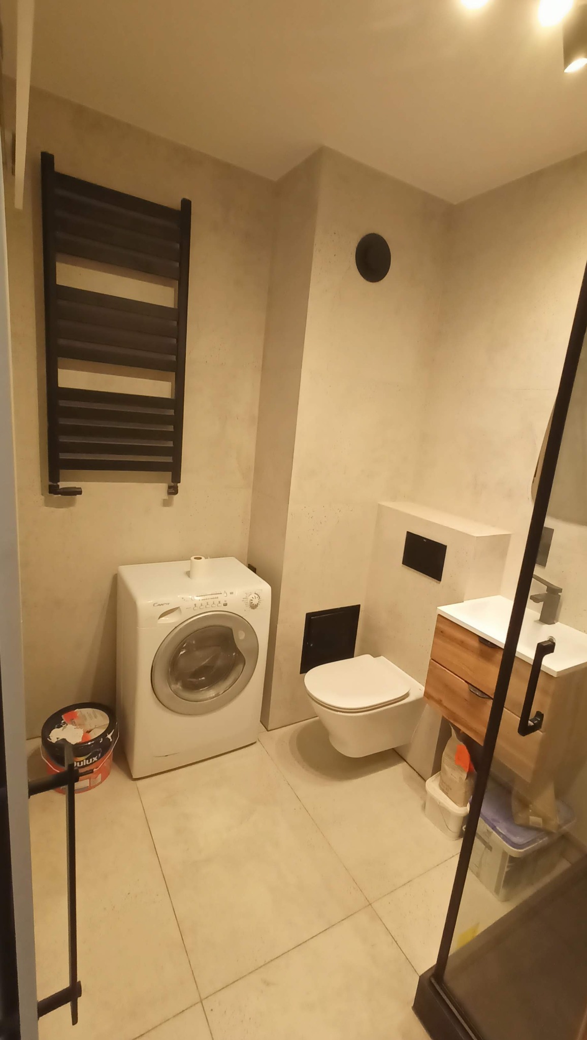 Jasna łazienka po remoncie: pralka, wisząca toaleta, umywalka z drewnianą szafką, grzejnik drabinkowy. Na podłodze płytki w odcieniach beżu. Widoczny fragment kabiny prysznicowej.