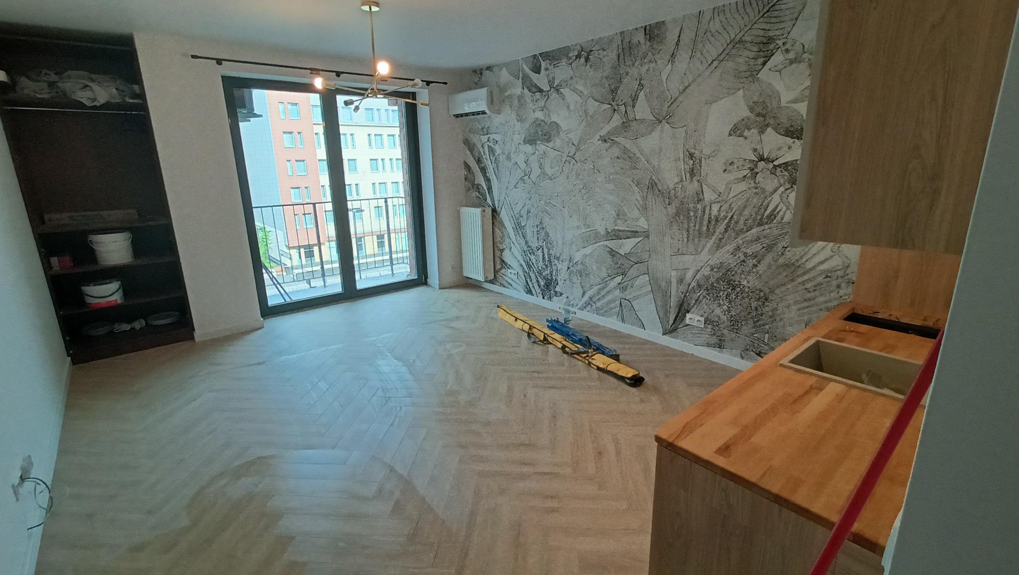 Jasne, przestronne wnętrze po remoncie: parkiet w jodełkę, fototapeta z motywem roślinnym, okno balkonowe z widokiem na miasto. Częściowo widoczna zabudowa kuchenna.